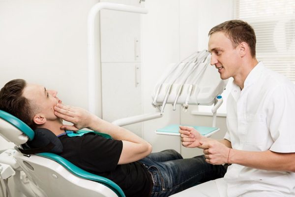 Young man sits in dentist’s chair holding one hand against his jaw, while a young dentist sits by his side speaking with him