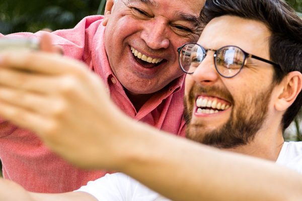 An older man and young man with glasses are smiling and laughing while taking a “selfie” photo on a smartphone