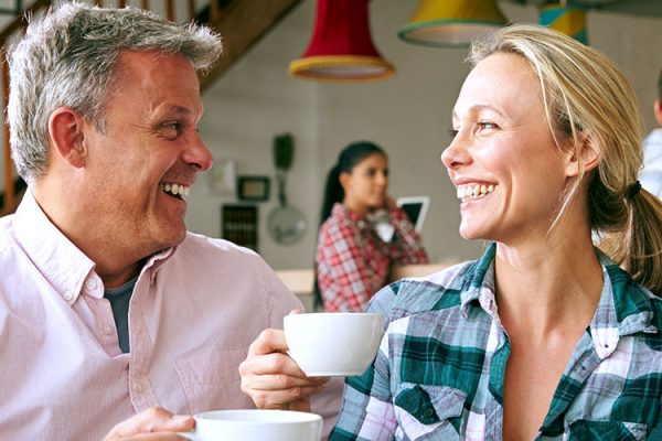 A man and woman in a coffee shop, sitting down and looking at each other while smiling and “cheers”-ing their coffee mugs