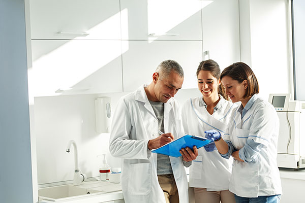 Implant dentist in coat lab holding a blue clipboard while consulting with two colleagues in dental office lab